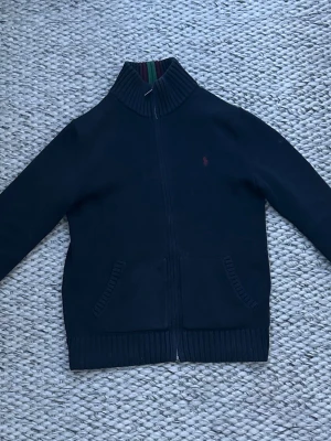 Mörkblå ziptröja från Polo Ralph Lauren - Mörkblå ziptröja från Polo Ralph Lauren med ribbad krage som har gröna och röda ränder. Tröjan har två fickor fram, diskret vinröd logga på bröstet och ribbade muddar vid ärmslut och nederkant. Barnstorlek L/G(14-16) motsvarar 164-170.
