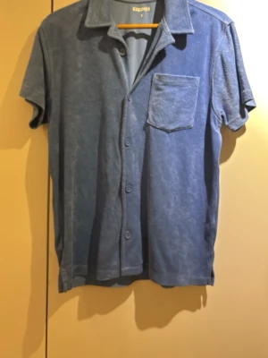 Blå kortärmad skjorta från Kappahl - Blå skjorta från Kappahl i mjukt frottématerial. Skjortan har korta ärmar, klassisk krage, bröstficka och knäppning framtill. Perfekt för en avslappnad och trendig look.