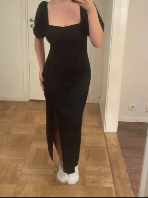Svart långklänning med puffärm - Stilren svart långklänning med fyrkantig urringning och puffärmar. Klänningen har en smickrande passform och en slits framtill som ger en trendig touch. Perfekt för dig som vill ha en elegant look med lite edge.