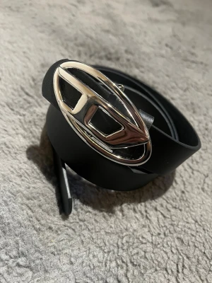 Svart bälte med silverfärgat spänne - Snyggt svart bälte med ett stort, ovalt silverfärgat spänne som har en unik design. Perfekt accessoar för att lyfta din outfit och ge en cool touch. Bältet är gjort i skinn och har en stilren look.