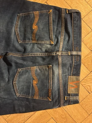 Mörkblå jeans från Nudie Jeans - Säljer ett par mörkblå jeans från Nudie Jeans med klassisk femficksmodell och orange kontrastsömmar. Jeansen har en snygg tvätt och Nudie-loggan på bakfickan samt patch i läder bak i midjan. Tillverkade i slitstark denim av bomull.