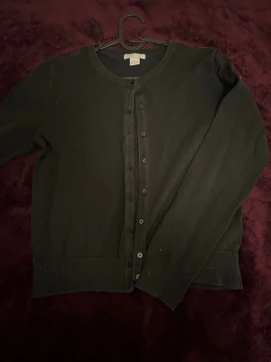 Svart cardigan med knappar - En klassisk svart cardigan med rund hals och knäppning framtill. Tröjan har långa ärmar och ribbade muddar vid ärmslut och nederkant. Perfekt att slänga över en t-shirt eller linne för en enkel och stilren look.