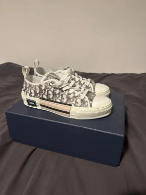 Dior B23 låga sneakers monogram - Säljer ett par Dior B23 låga sneakers i vitt och grått med det klassiska Dior-monogrammet på sidan. Skorna har genomskinliga paneler, vita snören och robust vit sula med logotypdetalj på sidan. Perfekta för dig som gillar exklusiv streetwear.
