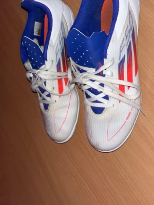 Adidas vita fotbollsskor med blå detaljer - Säljer ett par Adidas fotbollsskor i vitt med blå och röda detaljer. Skorna har snörning, låg profil och en ovandel i syntetmaterial. Insidan är blå med orange sula och yttersidan har grafiska ränder. Perfekta för dig som vill sticka ut på planen.