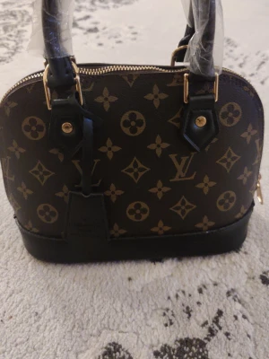 Louis Vuitton monogram handväska - Snygg handväska från Louis Vuitton med klassiskt brunt monogrammönster och svarta detaljer. Väskan har guldfärgade metalldetaljer och två handtag. Materialet är läder och väskan har en rundad form med dragkedja upptill.