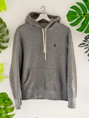 Grå hoodie från Polo Ralph Lauren - Snygg grå hoodie från Polo Ralph Lauren med vit dragsko och klassisk logga broderad på bröstet. Hoodien har känguruficka och är tillverkad i mjukt bomullsmaterial. Perfekt för en avslappnad och stilren look. Har ett litet märke vid ena handleden men som är svagt. Ingen annan defekt