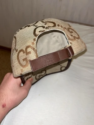 Beige GG keps från Gucci - Snygg beige keps från Gucci med klassiskt GG-mönster i brunt. Kepsen har justerbart brunt läderband bak och böjd skärm med läderdetaljer. Tillverkad i canvasmaterial med exklusiv känsla och tydlig logga både inuti och utanpå.