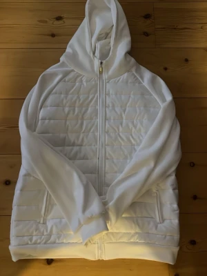 Vit cardigan med dragkedja - Snygg vit cardigan med quiltade paneler framtill och på ryggen. Dragkedja hela vägen, två fickor med dragkedja och en stor huva. Tillverkad i mjukt material med vadderad känsla, perfekt för kyliga dagar. Stilren och enkel design utan synligt märke.