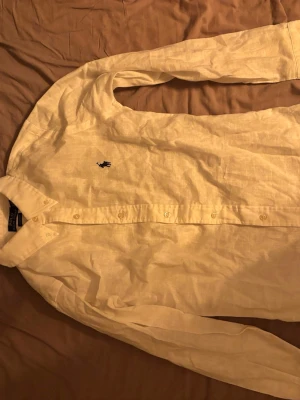 Vit linneskjorta från Ralph Lauren - Snygg vit skjorta i linne från Ralph Lauren med klassisk krage och lång ärm. Skjortan har knäppning framtill och den ikoniska broderade loggan på bröstet. Perfekt för en stilren och avslappnad look.