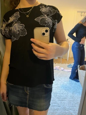 Svart blommig topp med vita detaljer - Snygg svart topp med vita blommiga linjemönster över axlar och bröst. Toppen har rund halsringning och korta ärmar. Perfekt till jeans eller kjol för en trendig look.