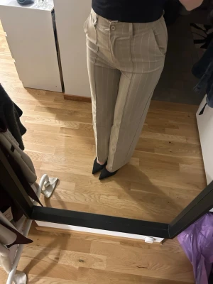 Beige kostymbyxor med vita ränder - Snygga beige kostymbyxor med raka ben och vita vertikala ränder. Byxorna har hög midja, fickor i sidorna och stängs med knapp och dragkedja. Perfekta för en stilren och trendig look.