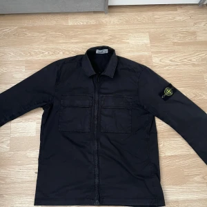 Svart overshirt från Stone Island - Snygg svart overshirt från Stone Island med dragkedja framtill och två stora bröstfickor. Klassisk krage och ikonisk logotyp-patch på vänster ärm. Perfekt för lager-på-lager och streetwear-stil.