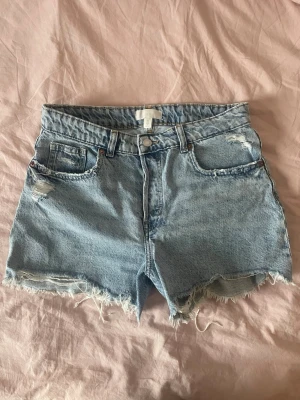 Ljusblåa distressed jeansshorts - Säljer ett par ljusblåa jeansshorts med hög midja och sliten, rå kant. Shortsen har slitna detaljer både fram och bak och knäpps åt med fem knappar. Perfekta för sommaren och ger en avslappnad vibe.