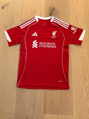Liverpool matchtröja Adidas röd - Röd Liverpool FC matchtröja från Adidas med vita detaljer och klubbmärke på bröstet. Tröjan har tryck från Standard Chartered och vita ränder på axlarna. Tillverkad i lätt och ventilerande material, perfekt för fotbollsträning eller match.