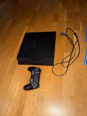 PlayStation 4 med handkontroll och spel - Säljer en svart PlayStation 4 med en trådlös DualShock 4-handkontroll och strömkabel. Konsolen är tillverkad i plast och har en stilren, fyrkantig design. Flera spel på Blu-ray medföljer, perfekt för gamingkvällar med vänner.