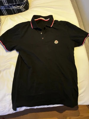 Svart pikétröja från Moncler - Svart pikétröja från Moncler med klassisk krage och knäppning framtill. Tröjan har röda och vita detaljer på krage och ärmslut samt Moncler-logga