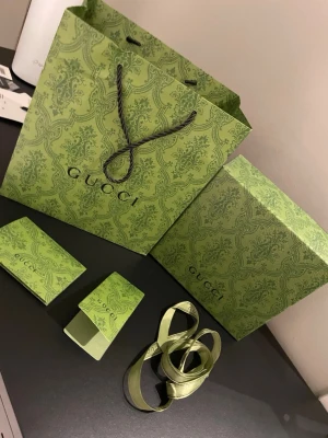 Gucci grön presentförpackning set - Snyggt set med Gucci presentpåse, ask, tygpåse, band och kort i matchande grön färg med elegant mönster. Perfekt för att ge bort eller förvara exklusiva accessoarer. Allt är dekorerat med klassiska Gucci-loggan och detaljer i premium papper och tyg.