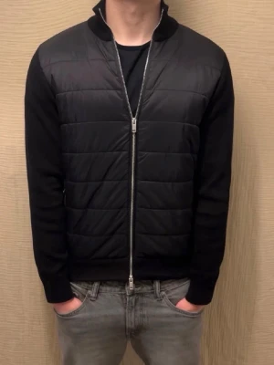 Svart quiltad cardigan med dragkedja - Snygg svart cardiganjacka med quiltad front och släta ärmar. Jackan har hög krage och dubbel dragkedja framtill. Perfekt för lager-på-lager och ger en clean look. Materialet ser ut att vara en mix av bomull och syntet.