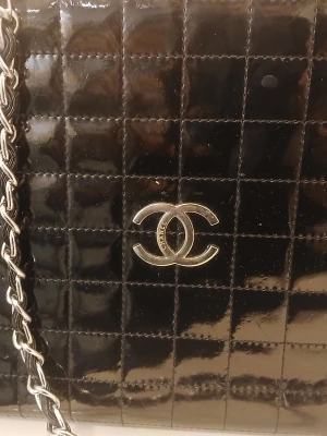Svart quiltad handväska från Chanel - Säljer en svart quiltad handväska från Chanel med klassiskt CC-logo i silver framtill. Väskan har en kedjerem i silverfärg och är tillverkad i glansigt läder. Perfekt för dig som vill ha en ikonisk och tidlös accessoar med lyxig känsla.