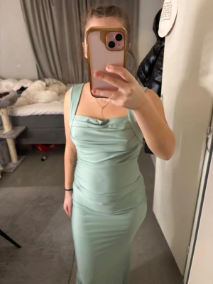 Ljusgrön långklänning med drapering - Snygg ljusgrön långklänning med breda axelband och draperad detalj framtill. Klänningen har en figurnära passform och är tillverkad i ett mjukt, stretchigt material som ger en smickrande siluett. Perfekt för dig som vill sticka ut med en elegant look.