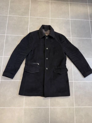 Svart ullkappa från Oscar Jacobson - Stilren svart kappa från Oscar Jacobson i ullmix med klassisk krage, knäppning framtill och två stora fickor med lock samt en ficka med dragkedja. Perfekt för dig som vill ha en tidlös och elegant look till hösten eller vintern.