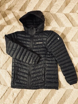 JR FROST DOWN HOOD JACKET  - Mörkblå lättviktsdunjacka med huva från Peak Performance. Innehållet i jackan är 90% ankdun och 10% fjäder. Märkets logga är broderad och placerad på bröstet och på ärmen. Jackan har två fickor framtill som stängs med dragkedja. Avtagbar huva.