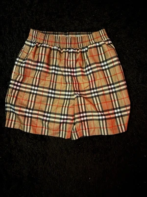 Rutig Burberry shorts i beige - Säljer ett par klassiska rutiga shorts från Burberry i beige med svarta, vita och röda detaljer. Shortsen har elastisk midja och sidofickor. Tillverkade i ett lätt och bekvämt material, perfekta för sommaren.