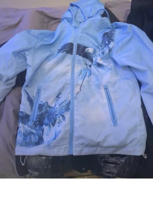 Blå hoodie med örntryck från Trapstar - Säljer en ljusblå hoodie från Trapstar med dragkedja och huva. Hoodien har stora, detaljerade örntryck både fram och bak samt Trapstar-loggan i vitt på ryggen. Två fickor med dragkedja framtill. Perfekt för dig som gillar streetwear och unika prints. Priset Kan diskuteras vid snabbt äffar mvh😁