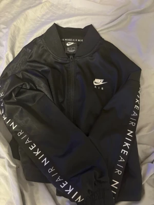 Svart Nike Air zip hoodie - Svart zip hoodie från Nike Air med vit logga på bröstet och tryckt text 'NIKEAIR' längs båda ärmarna. Tröjan har hög krage, dragkedja framtill och är tillverkad i ett mjukt material som känns skönt mot huden. Perfekt för en sportig och avslappnad stil.