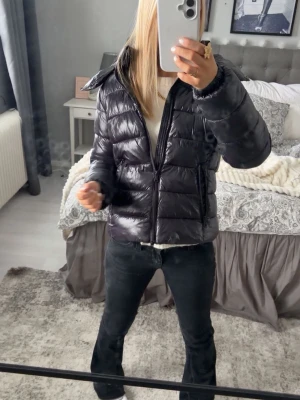 Svart glansig pufferjacka - Säljer en svart, glansig pufferjacka med hög krage och dragkedja framtill. Jackan har två sidofickor med dragkedja och en kortare, boxig passform som ger en trendig look. Perfekt för kalla dagar när du vill hålla värmen och ändå se snygg ut.
