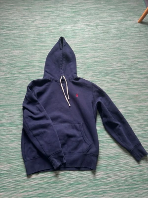 Mörkblå hoodie från Polo Ralph Lauren - Klassisk mörkblå hoodie från Polo Ralph Lauren med röd broderad logga på bröstet och vit snörning i huvan. Hoodien har känguruficka och långa ärmar. Perfekt för en avslappnad och stilren look.passad dig runt 170 cm