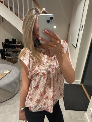 Blommig blus från Neo Noir - Söt blommig blus från Neo Noir i ljusrosa nyans med röda och gröna blommor. Modellen har volangdetaljer vid axlarna, v-ringning och är ärmlös. Luftigt material som passar perfekt till varma dagar. Märkt med 'Jayla Dreamy Rose Top'.