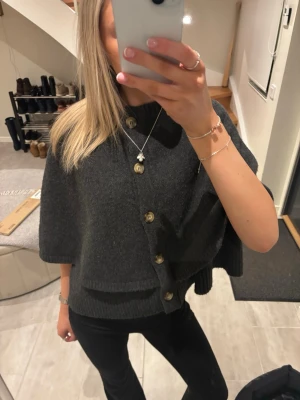 Mörkgrå ponchokofta med knappar - Ponchokofta i mjuk mörkgrå ullmix med stora, glansiga knappar framtill. Modellen är vid och har korta ärmar, vilket ger en avslappnad och trendig look. Perfekt att slänga över en topp för extra värme och stil.