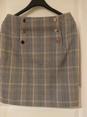 Rutig kortkjol med knappar - Snygg rutig kortkjol i grått och beige med dekorativa knappar framtill. Kjolen har en klassisk rak passform och är perfekt för dig som gillar en stilren look. Tillverkad i ett mjukt material som känns bekvämt hela dagen.