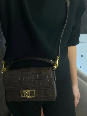 Mörkbrun RL handväska i croc-mönster - Brun handväska från Ralph Lauren i crokodil läder. Väskan har guldfärgade detaljer, RL-logga på spännet och både axelrem och handtag. Helt oanvänd så väskan är i nyskick. 