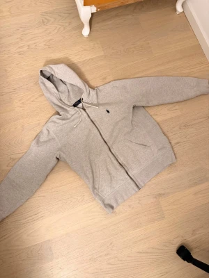Grå hoodie från Polo Ralph Lauren ÄKTAA!! - Snygg ljusgrå hoodie från Polo Ralph Lauren med dragkedja framtill och klassisk mörkblå logga broderad på bröstet. Hoodien har huva med vita dragsnören och två fickor framtill. Tillverkad i mjuk bomullsblandning för extra komfort. Pris kan diskuteras!
