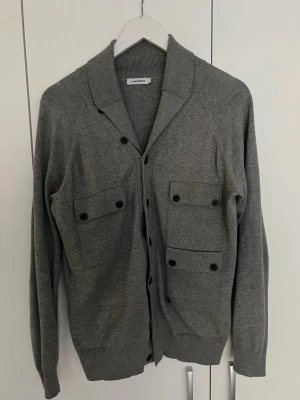 Grå cardigan från J. Lindeberg - Stilren grå cardigan med knäppning framtill från J. Lindeberg. Tröjan har två stora bröstfickor med lock och knappar samt ribbade muddar och krage. Perfekt för lager-på-lager och enkel att matcha med andra plagg.