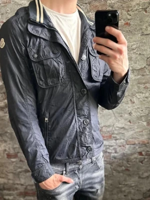 Mörkblå vindjacka från Moncler - Snygg mörkblå vindjacka/fieldjacket från Moncler med klassisk krage och vita ränder. Jackan har flera fickor med knappar och dragkedja, samt stora knappar framtill. Perfekt för dig som vill ha en stilren look.