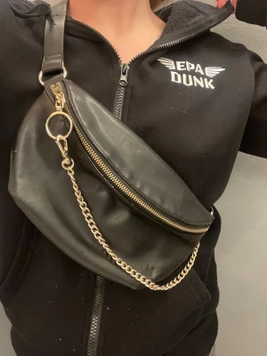 Svart axelväska med kedjedetalj - Snygg svart axelväska/magväska i slätt konstläder med justerbart axelband och guldfärgad kedjedetalj. Väskan har en dragkedja upptill och en rund ring som extra detalj. Perfekt för att lyfta din outfit med en trendig touch. Vill bli av med. Ej bra pris,,,, skriv till mig