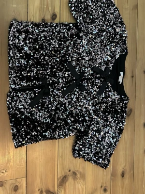 Svart paljettkofta med rosetter - En festlig, glittrig svart kofta täckt av silvriga paljetter och dekorerad med tre svarta rosetter framtill. Kort ärm och rund halsringning. Perfekt för dig som vill sticka ut och älskar detaljer. 
