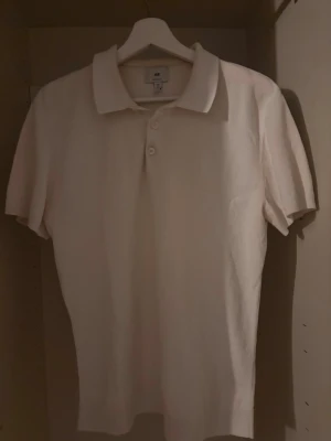 Vit pikétröja från H&M - Klassisk vit pikétröja från H&M med korta ärmar och krage. Tröjan har två knappar framtill och är tillverkad i mjukt material som känns skönt mot huden. Enkel och stilren design som passar till mycket.