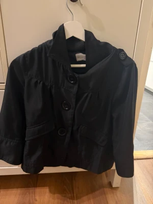 Svart kort rock med stora knappar - Modellen är trekvartsärmsd och är kortare med två fickor framtill och har en hög krage. 💞