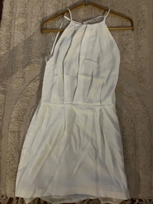 Vit kort klänning med spetsdetalj - Säljer en vit kort klänning med tunna axelband och hög halsringning. Baksidan har en öppen rygg med vacker spetsdetalj upptill. Klänningen har en figurnära passform och är perfekt för sommarens alla tillfällen.