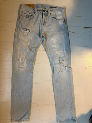 Dondup Jeans - Säljer ett par ljusblå jeans från Dondup med snygga slitningar och hål på både fram- och baksidan. Jeansen har klassisk femficksdesign, raka ben och är tillverkade i denim. Perfekta för en avslappnad och trendig stil.