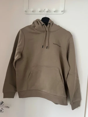 Beige hoodie från Peak Performance - Snygg beige hoodie från Peak Performance med broderad logga på bröstet. Hoodien har en stor känguruficka framtill, dragsko i huvan och ribbade muddar vid ärmslut och nederkant. Tillverkad i mjuk bomullsblandning för extra komfort. Aldrig använd, nyskick utan prislapp. 