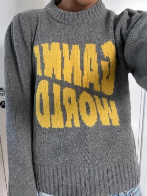 Grå stickad tröja Ganni World - Grå stickad tröja från Ganni med gul text 'GANNI WORLD' framtill. Tröjan har rund halsringning, ribbade muddar och en avslappnad passform. Perfekt för dig som gillar streetstyle och vill ha något bekvämt men ändå trendigt.