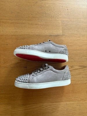 Beiga Louboutin sneakers med nitar - Säljer ett par beiga sneakers från Christian Louboutin i mocka med ikoniska röda sulor. Skorna har coola silverfärgade nitar på tå och häl, samt vita sulor. Perfekta för dig som vill sticka ut med en edgy och lyxig stil.
