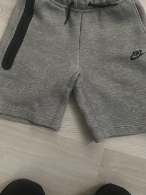 Grå Nike shorts med dragkedja 147-158 - Grå shorts från Nike med svart logga på vänster ben. Shortsen har snörning i midjan och en ficka med svart dragkedja på höger sida. Tillverkade i mjukt syntetmaterial som passar perfekt för träning eller chill.