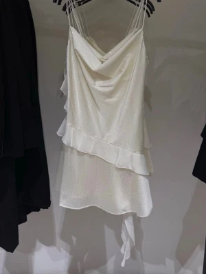 Vit kort klänning med volang ZARA - Säljer en vit kort klänning med tunna axelband och draperad urringning. Klänningen har glittrigt tyg, volangdetaljer och en lätt asymmetrisk nederkant. Perfekt för dig som vill ha en söt och trendig look. 