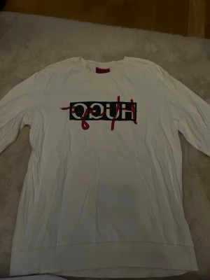 Vit långärmad HUGO tröja - Säljer en vit långärmad tröja från HUGO med en stor logga i svart och rött framtill. Tröjan har en klassisk rund halsringning och är gjord i mjuk bomull. Perfekt för dig som gillar streetwear och vill sticka ut med en snygg logga.